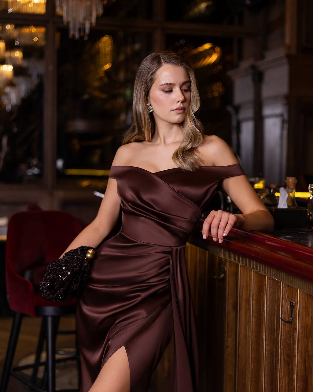 Woman in a brown off-shoulder dress leaning against a bar counter in a dimly lit room.