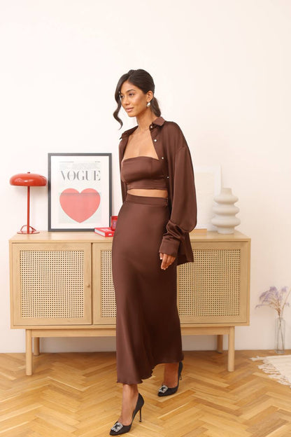 Woman in a brown outfit standing in a room with a wooden cabinet and decorative items.