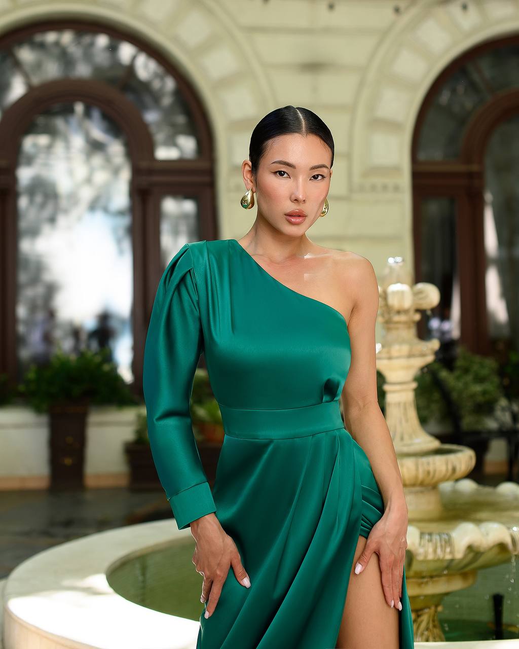 Woman in a green one-shoulder dress standing in an elegant outdoor setting with a fountain.