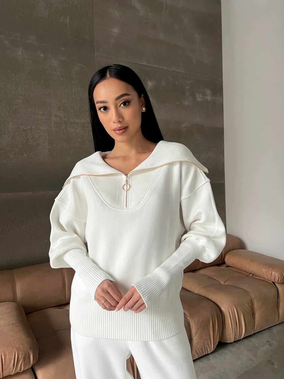 Woman wearing a white sweater and pants standing in a room with a brown sofa.
