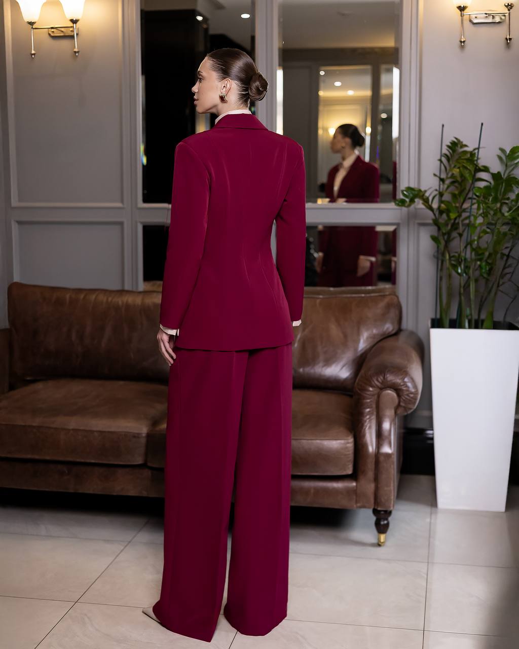 Woman in a burgundy suit standing in a room with a brown leather couch and a plant.