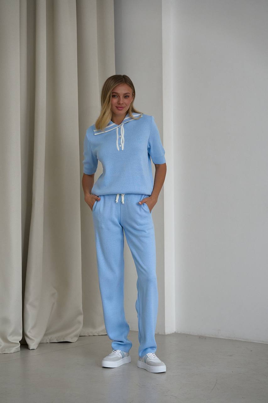Woman wearing a light blue tracksuit standing against a plain wall with a curtain.