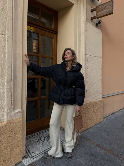 Woman in a black puffer jacket and white pants standing on a street corner.