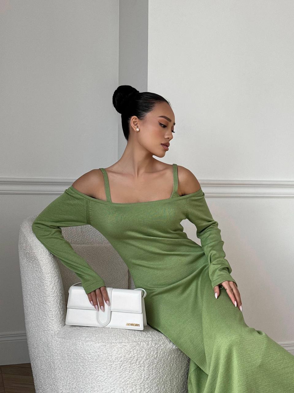 Woman in a green dress sitting on a white chair holding a white handbag.