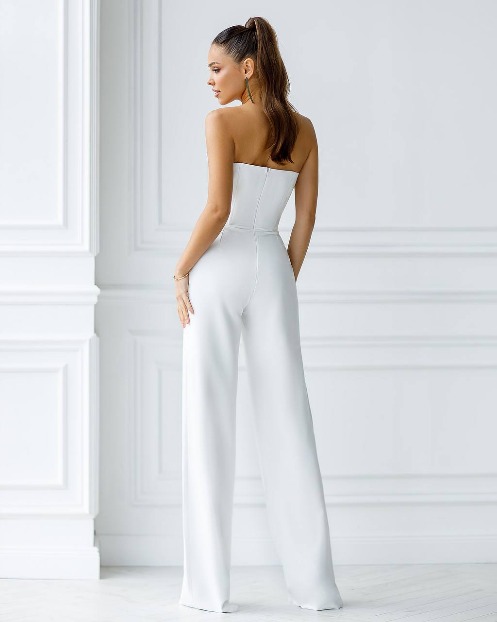 Woman wearing a white strapless jumpsuit against a light-colored wall.