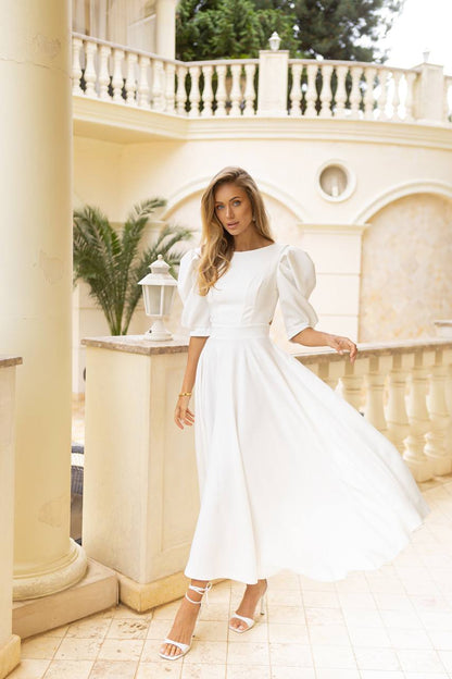 Woman in a white dress standing in an elegant outdoor setting with columns and plants.