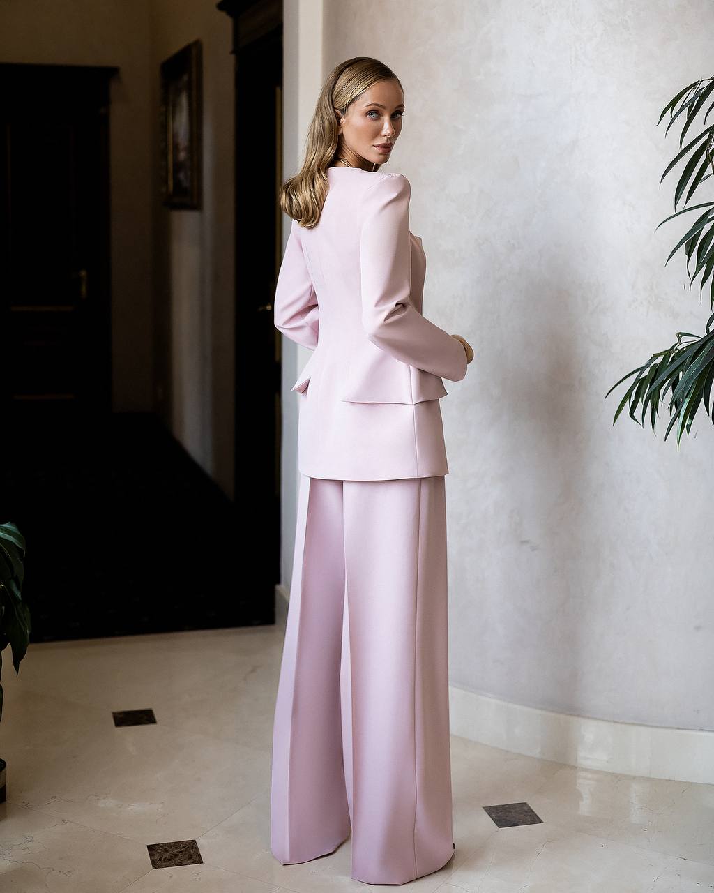 Woman in a light pink suit standing in a room with a plant in the corner.