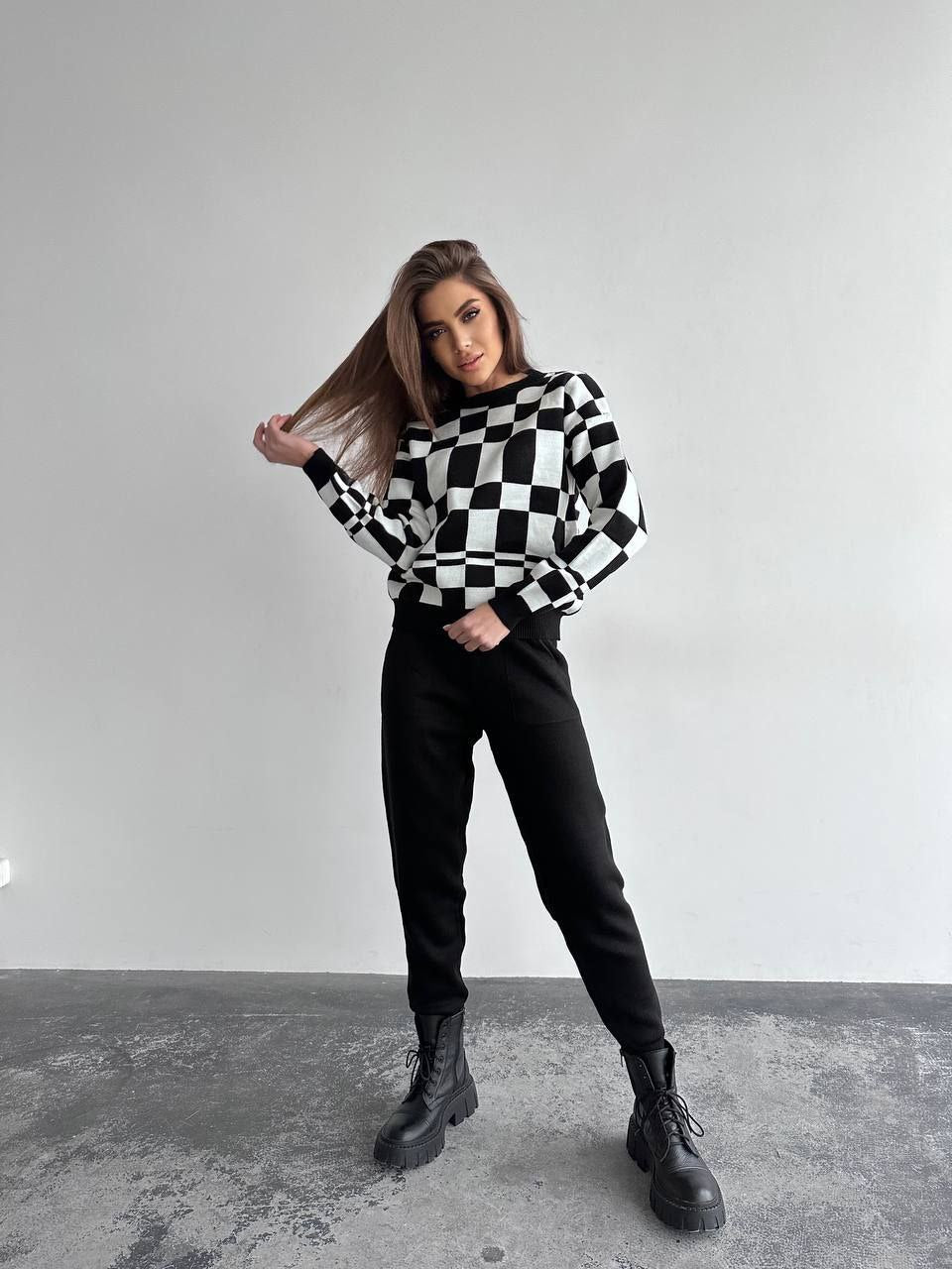 Woman wearing a black and white checkered sweater with black pants and boots on a plain background