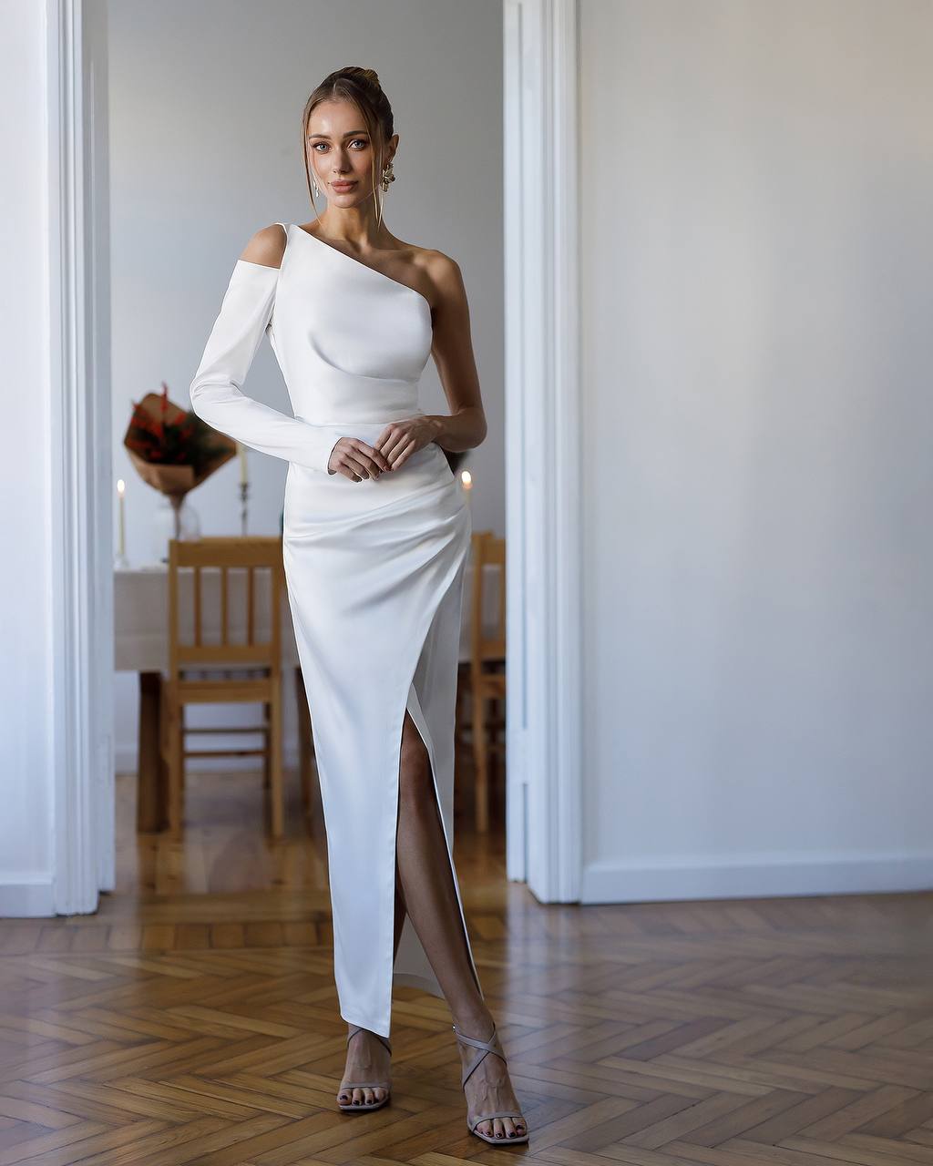 Woman in a white one-shoulder dress standing in a room with wooden flooring.
