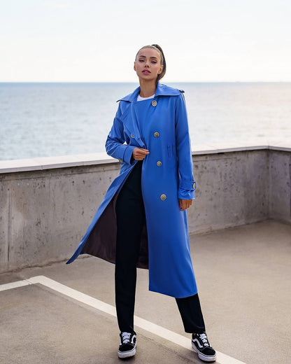 Person wearing a blue trench coat standing on a rooftop with a body of water in the background