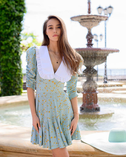 Woman in a light blue dress with a white collar standing in front of a fountain.