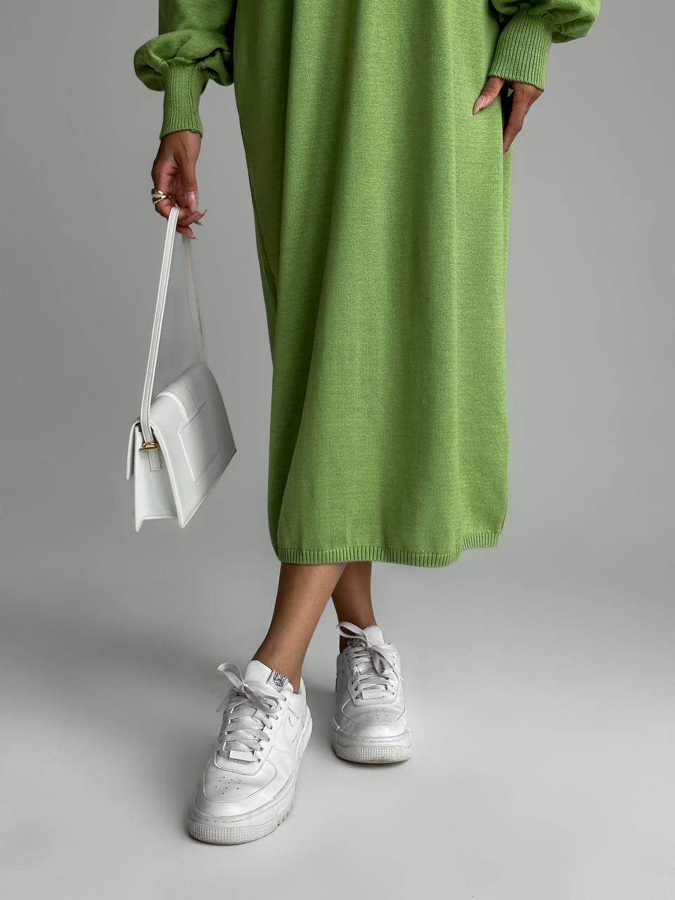 Person wearing a green dress and white sneakers holding a white bag on a gray background