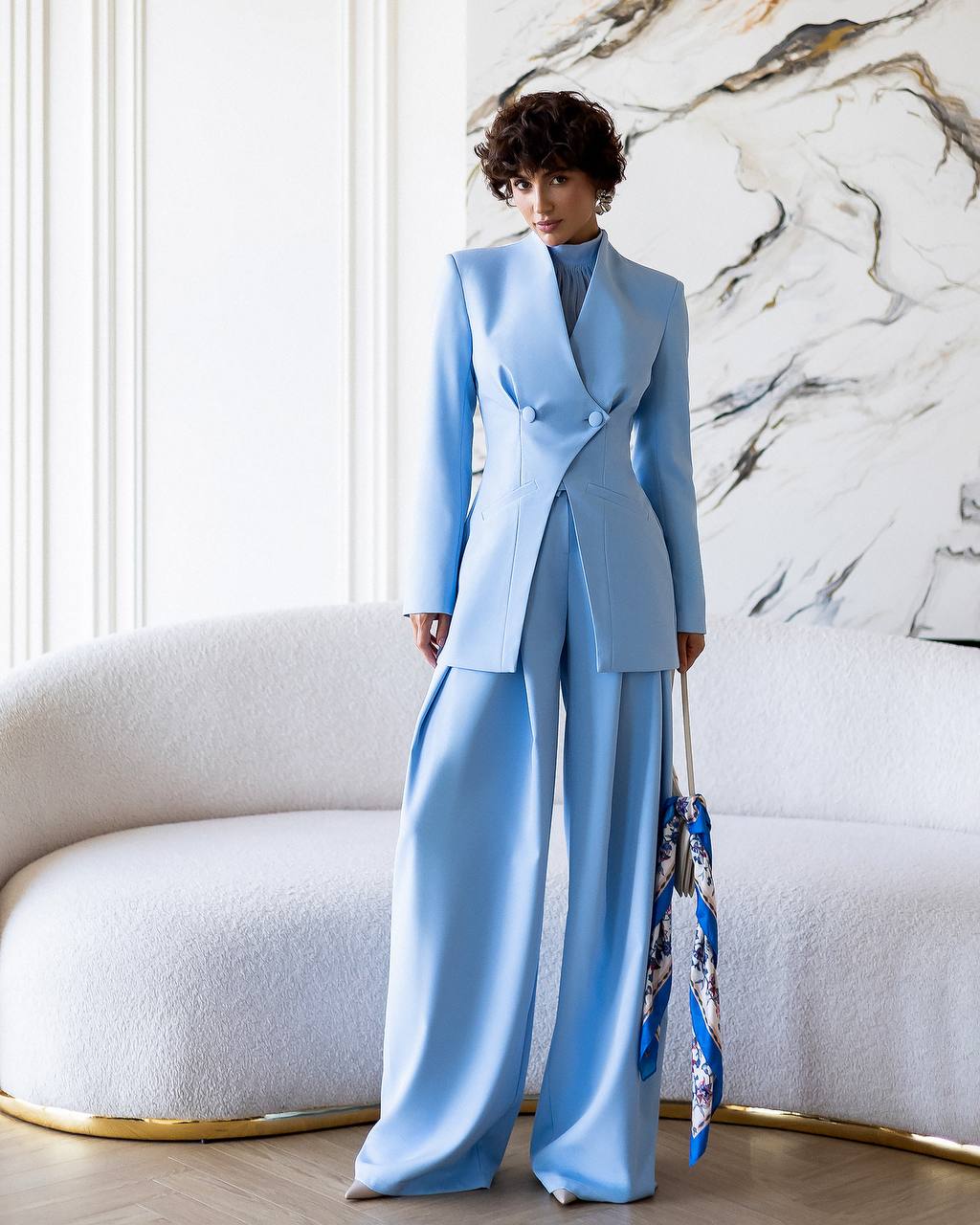Person wearing a light blue suit standing in a stylish room with marble wall.