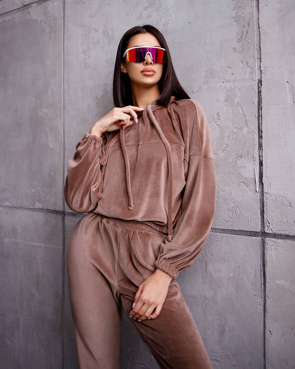 Person wearing a brown tracksuit with reflective sunglasses against a gray wall.