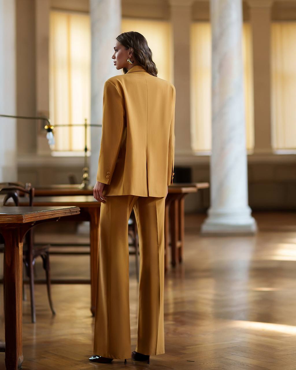Woman in a mustard yellow suit standing in a room with wooden tables and columns.