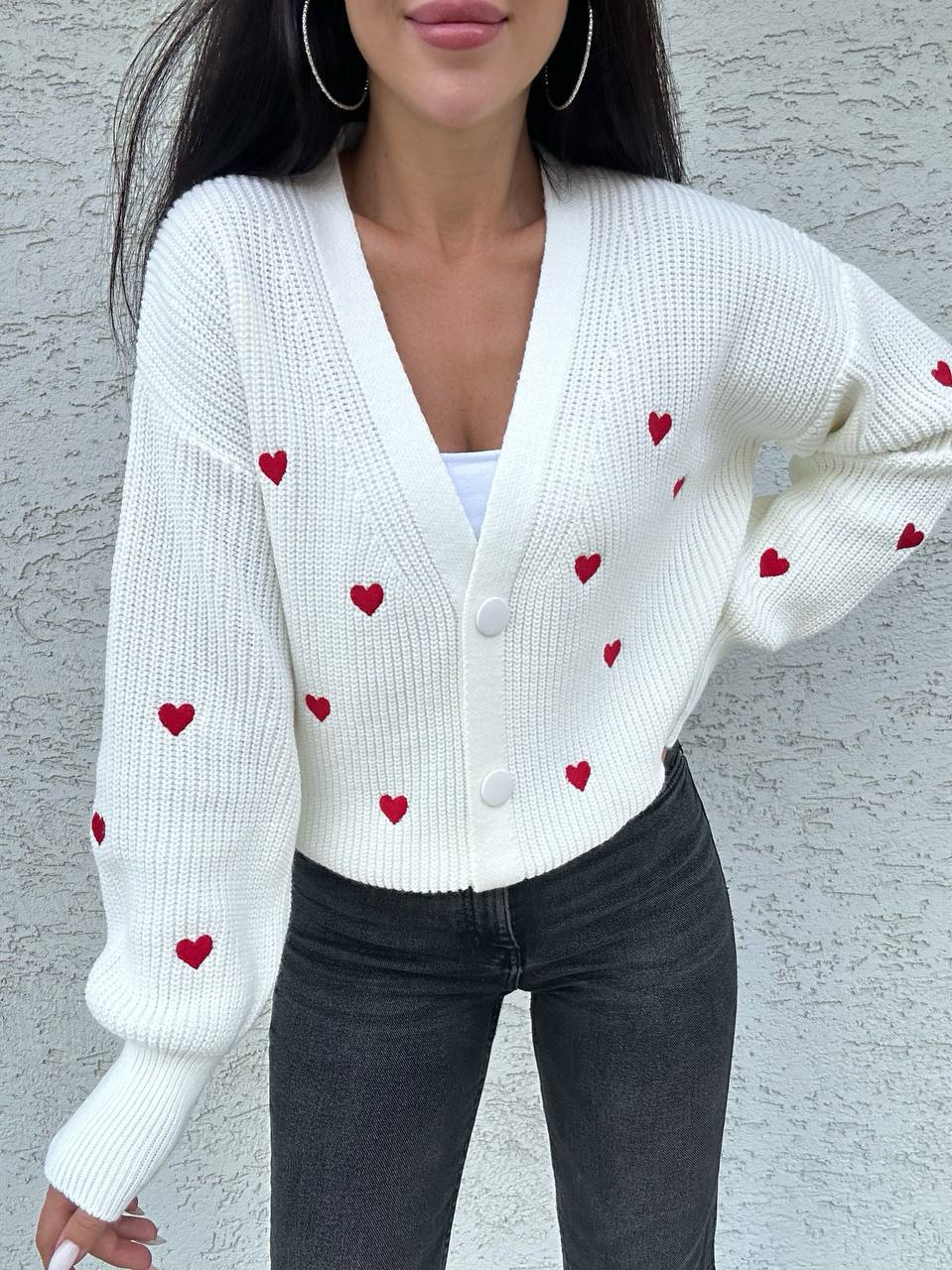 White cardigan with red heart patterns worn by a person against a gray background