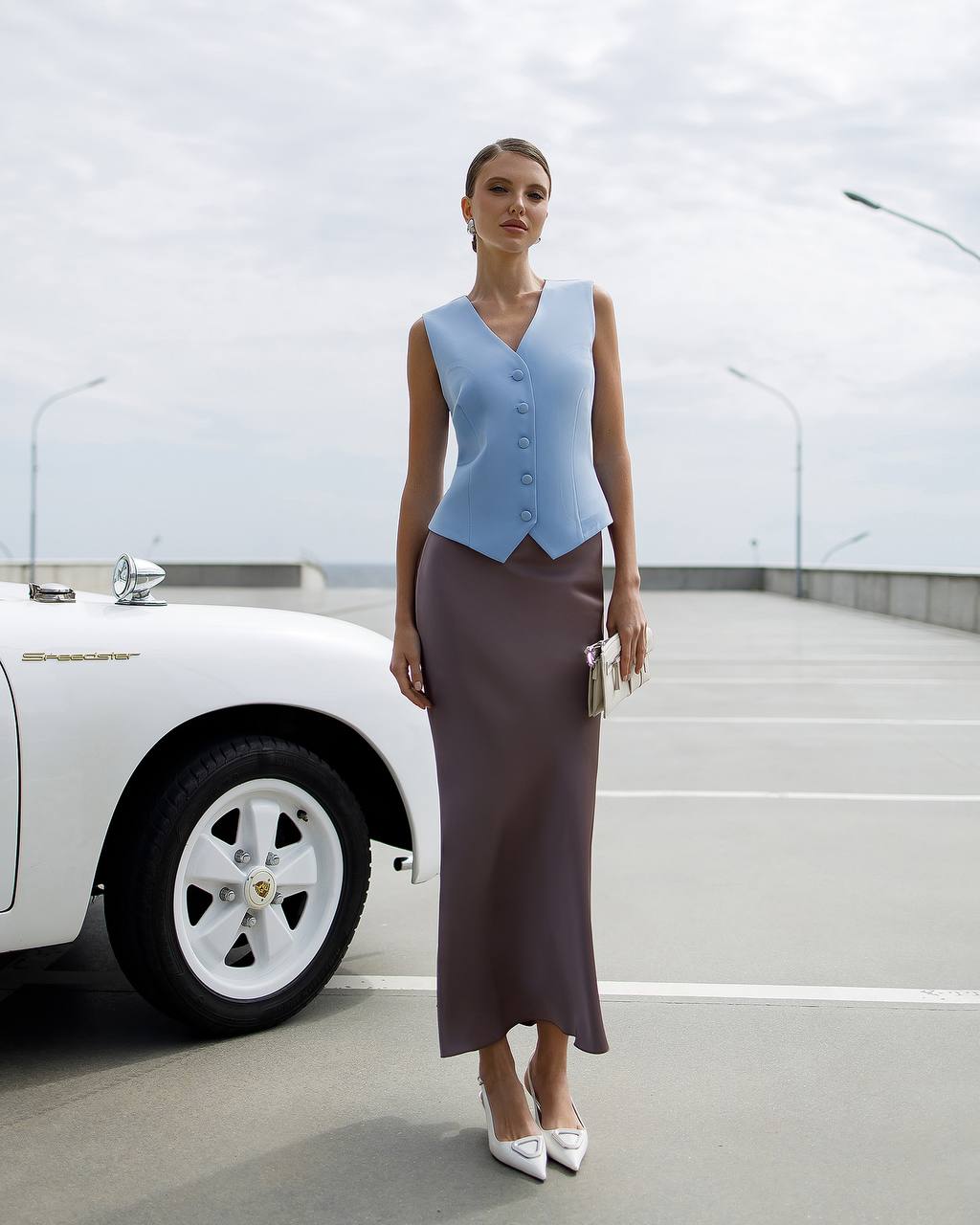 Woman in a light blue sleeveless top and brown skirt standing next to a white car on a road.