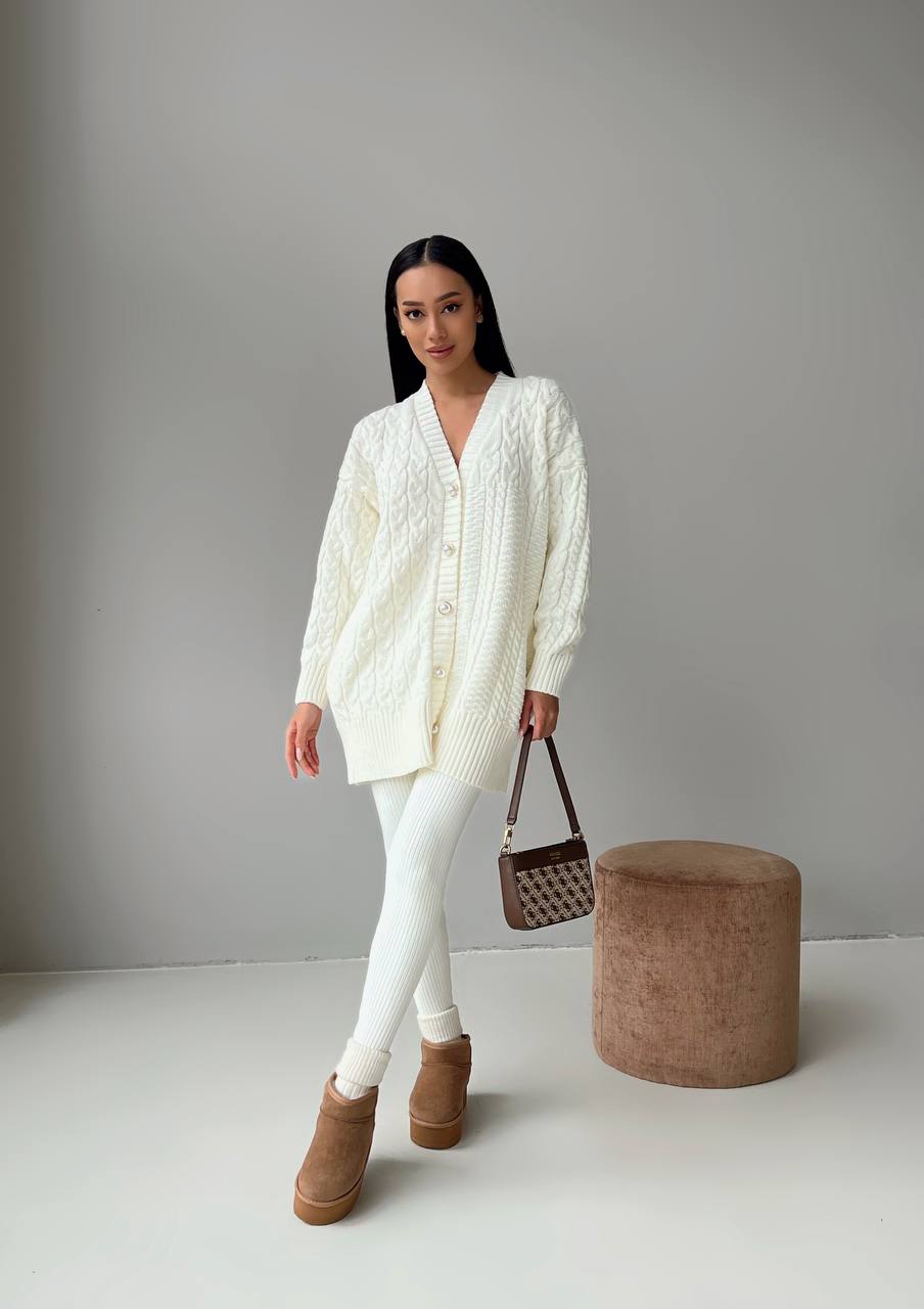 Woman wearing a white cable knit cardigan, white pants, and brown boots, standing next to a brown bag and ottoman.