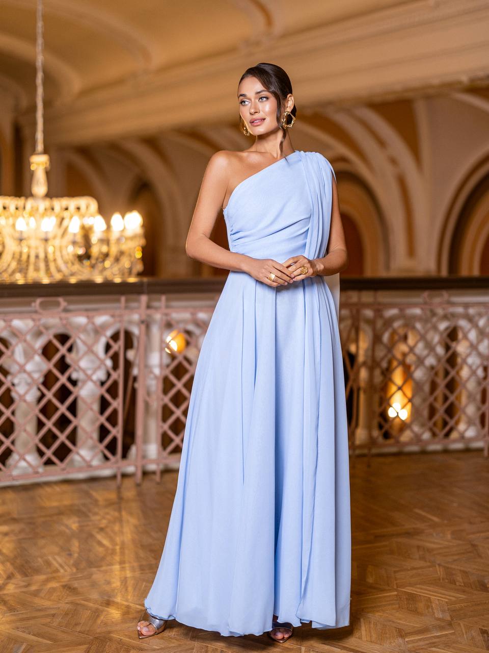 Woman in a light blue one-shoulder gown standing in an elegant indoor setting with chandeliers.