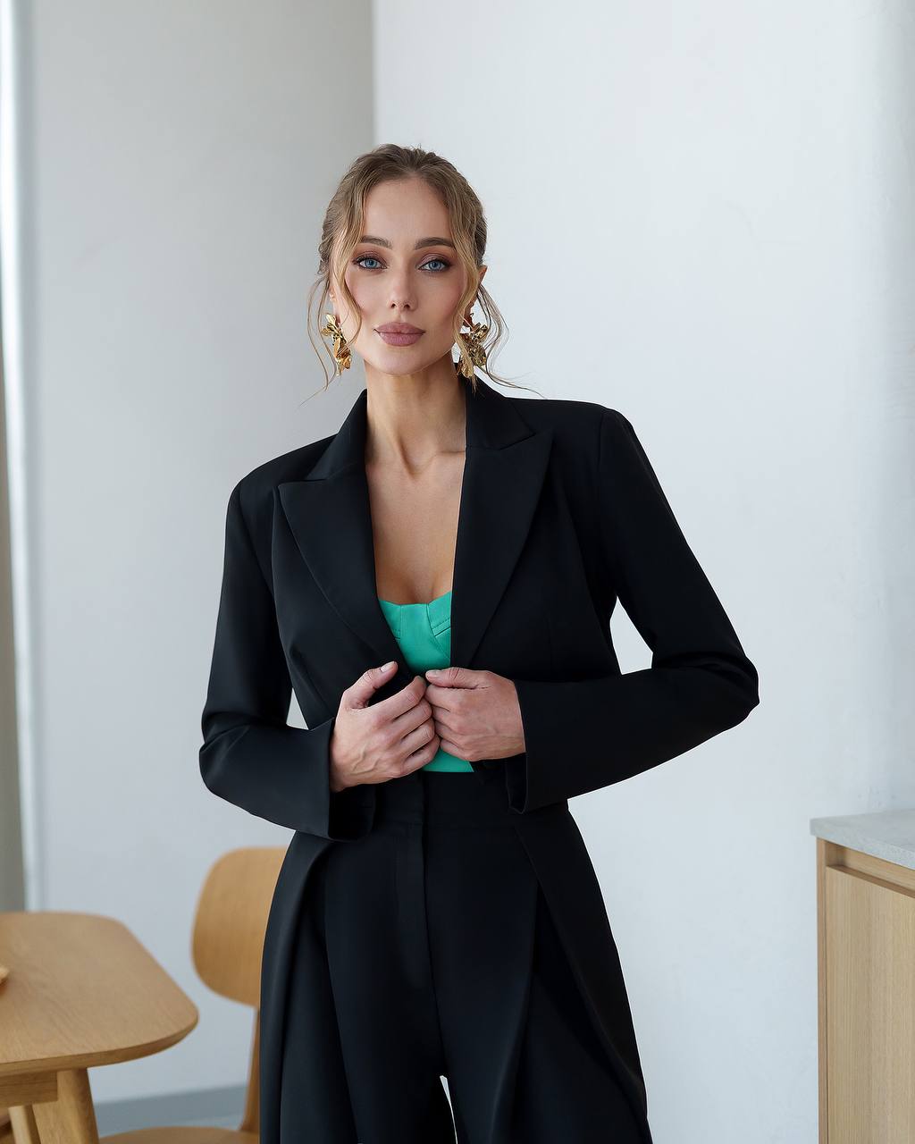 Woman wearing a black blazer over a green top in a minimalistic room.