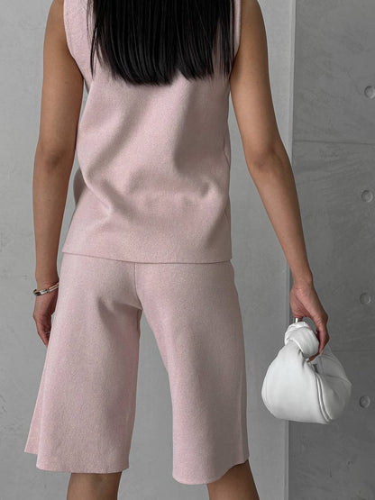 Person wearing a pink sleeveless top and shorts set, holding a white handbag against a gray background.