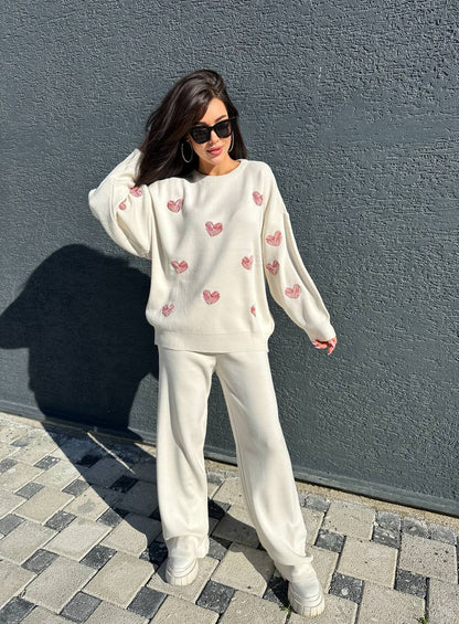 Woman wearing a white sweatshirt with pink heart patterns and white pants against a gray wall.