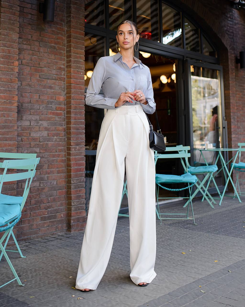 Woman in light gray shirt and white wide-leg pants standing outside a brick building with teal chairs.