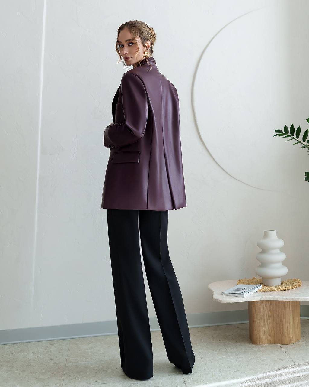 Woman in a purple coat and black pants standing in a modern interior setting.