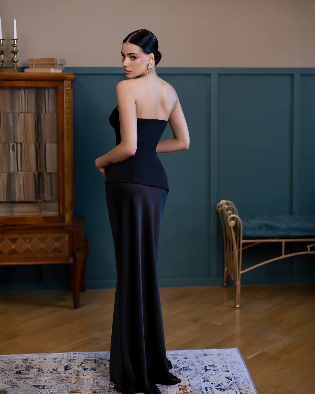 A woman stands in a formal indoor room wearing a strapless black gown with a fitted corset-style bodice and a long flowing skirt. The room has wood paneling, a wooden cabinet, a bench, and a patterned rug.
