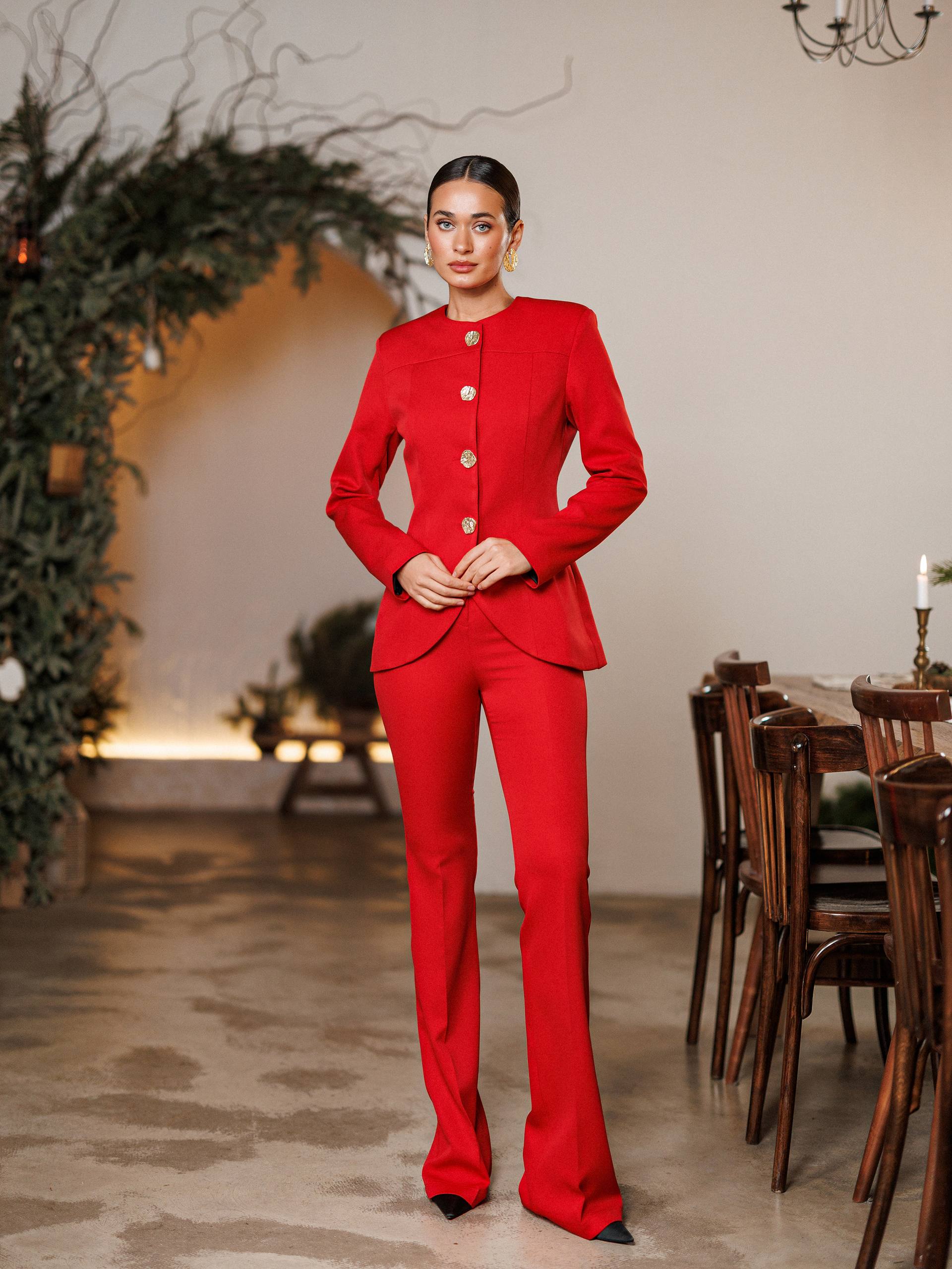 Woman in a red suit standing in an elegant room with a chandelier and wooden furniture.