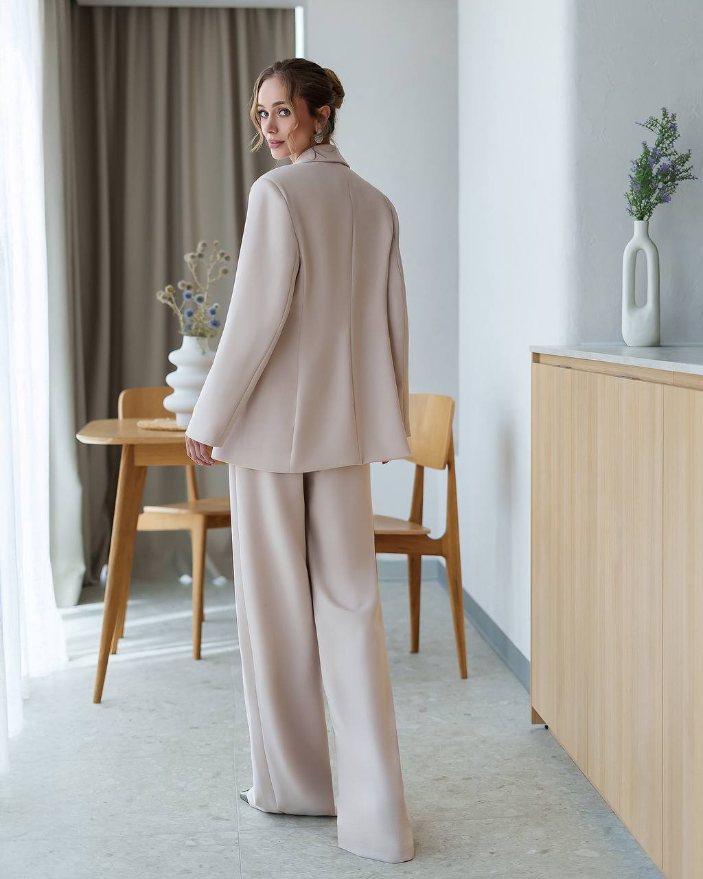 Woman in a beige suit standing in a modern room with wooden furniture and decor.