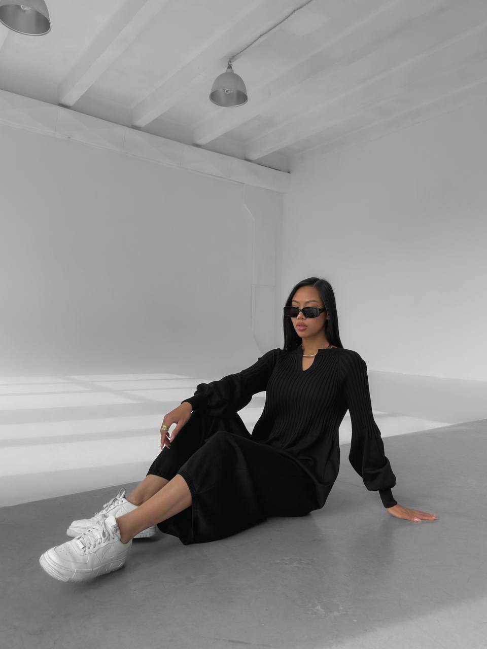 Woman in a black dress and white sneakers sitting on a gray floor in a minimalistic room.