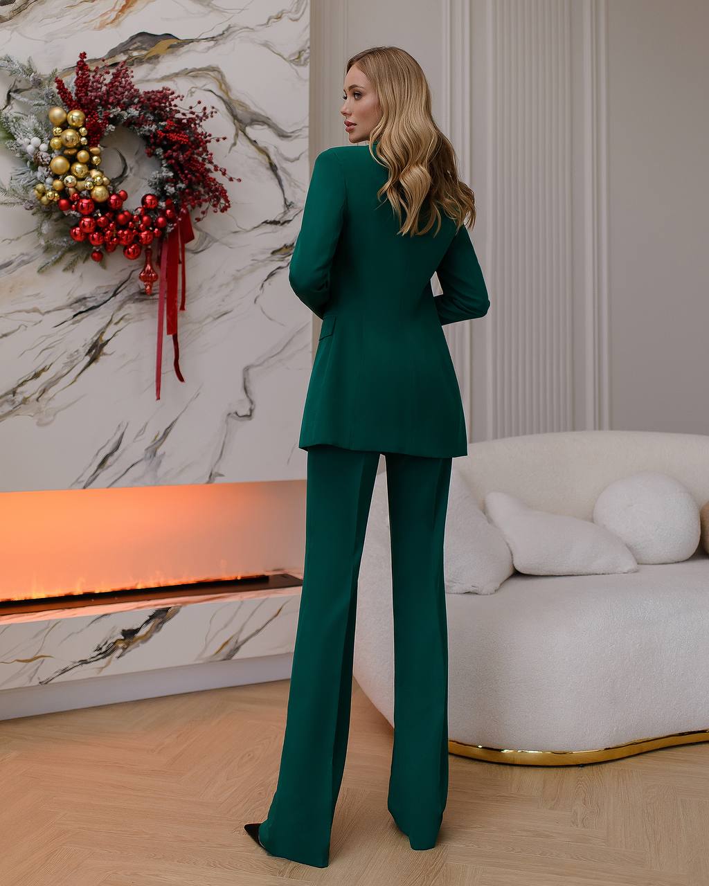 Woman in a green suit standing in a stylish room with a decorative wreath on the wall.