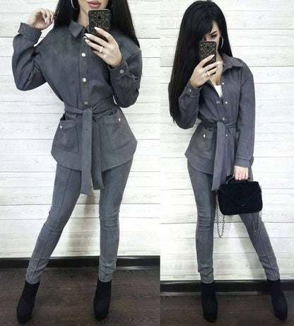 Woman in a gray outfit with a belt, holding a black clutch against a white wall.