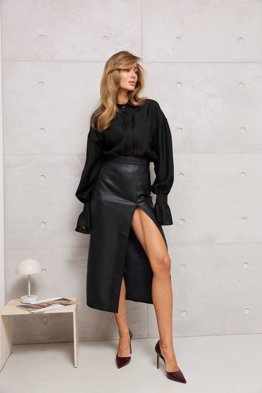 Woman wearing a black outfit with a high slit skirt against a light gray wall.