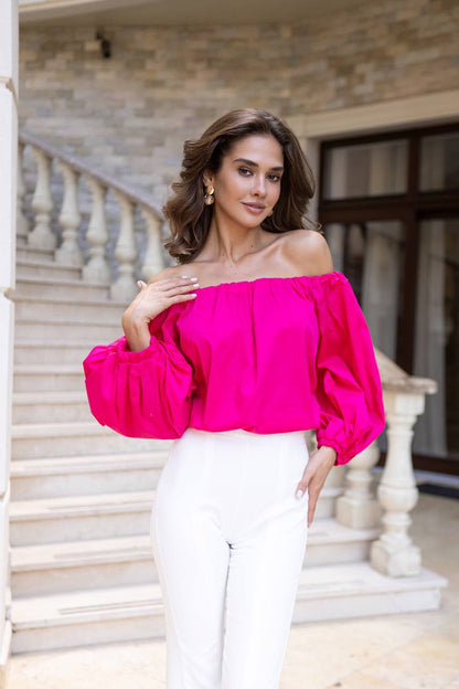 Woman wearing a bright pink off-shoulder top and white pants in an elegant interior setting.