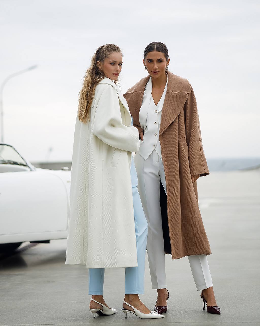 Two women in stylish outfits standing next to a white car with a blurred background