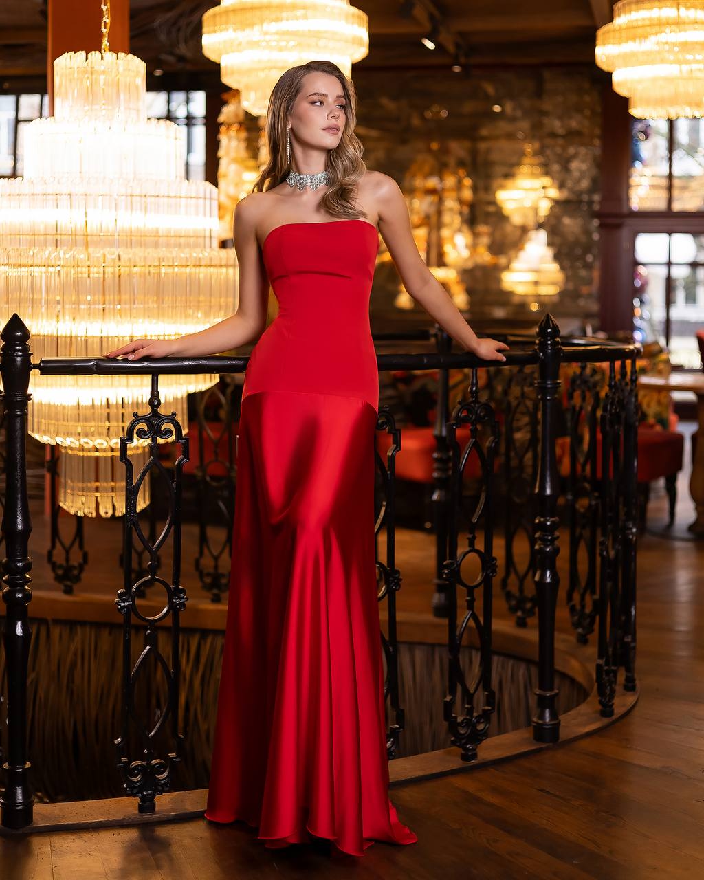 Woman in a red strapless gown standing in an elegant interior setting with chandeliers.