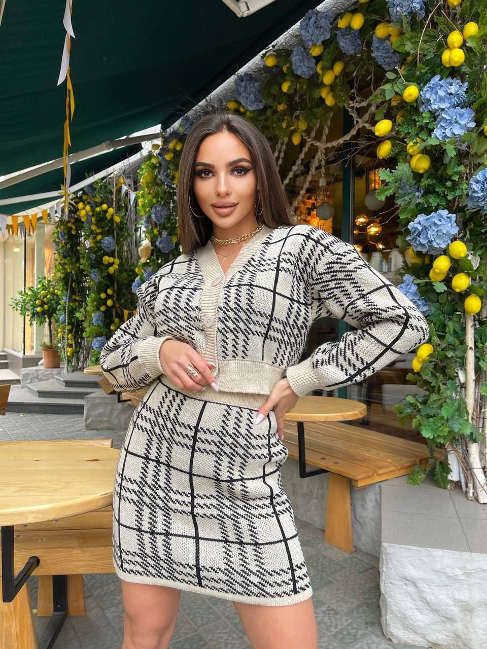 Woman in a black and white checkered outfit standing in an outdoor setting with decorative plants and tables.