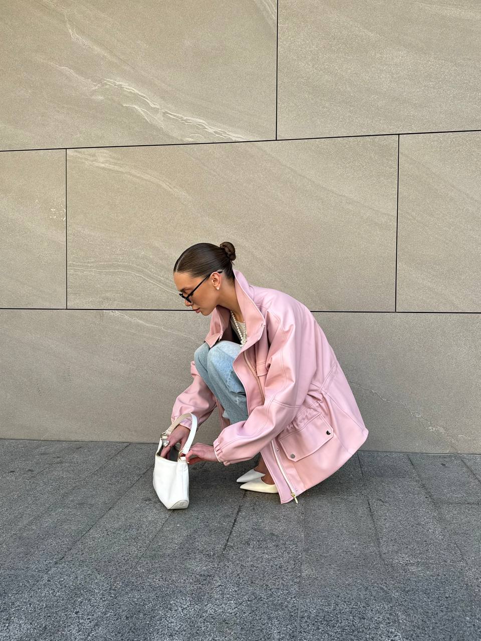 Person wearing a pink jacket and light blue jeans, crouching against a gray wall.