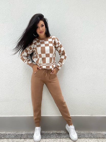 Woman wearing a checkered sweater and brown pants against a light gray wall.