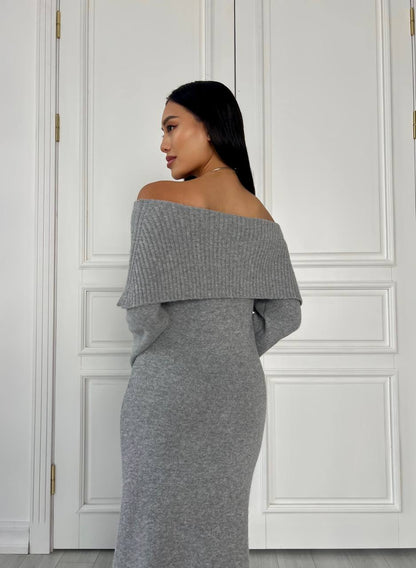 Woman wearing a gray off-shoulder dress against a white paneled wall.