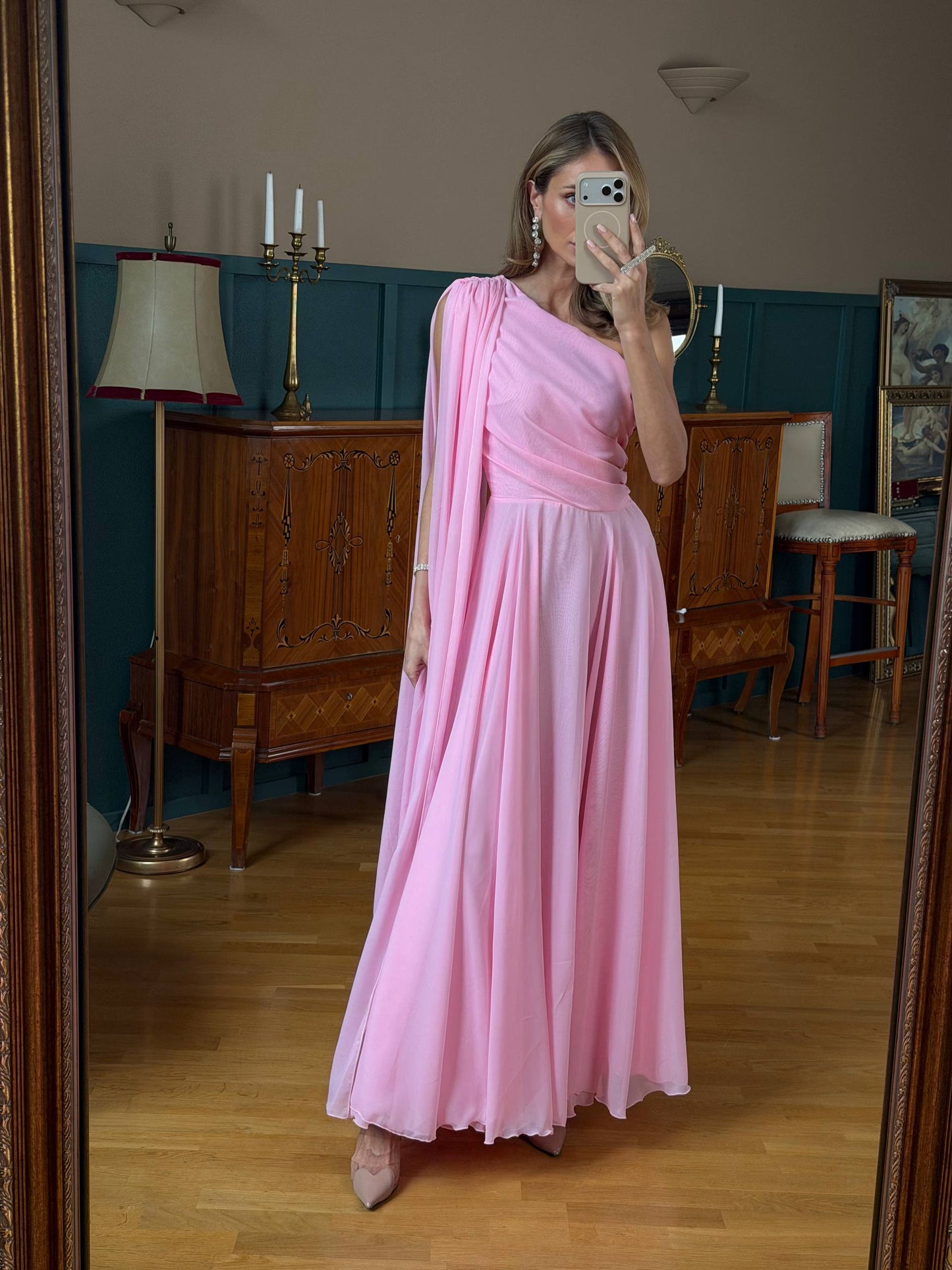 Woman in a pink dress taking a mirror selfie in a room with wooden furniture and a lamp.