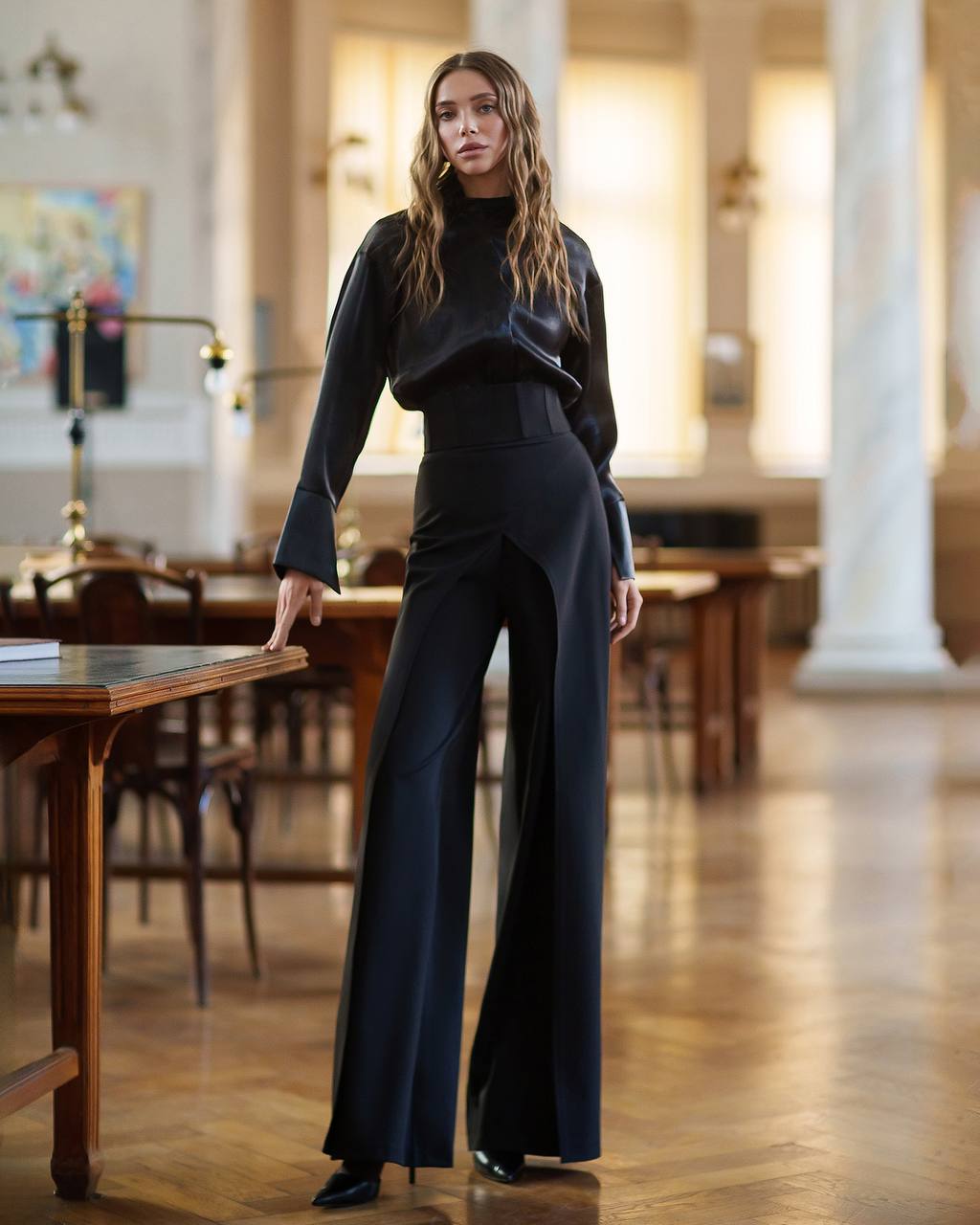 Woman in a black outfit standing in an elegant room with wooden floors and columns.