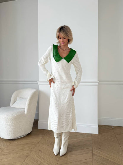Woman wearing a white dress with a green collar in a room with a white wall and a white chair.