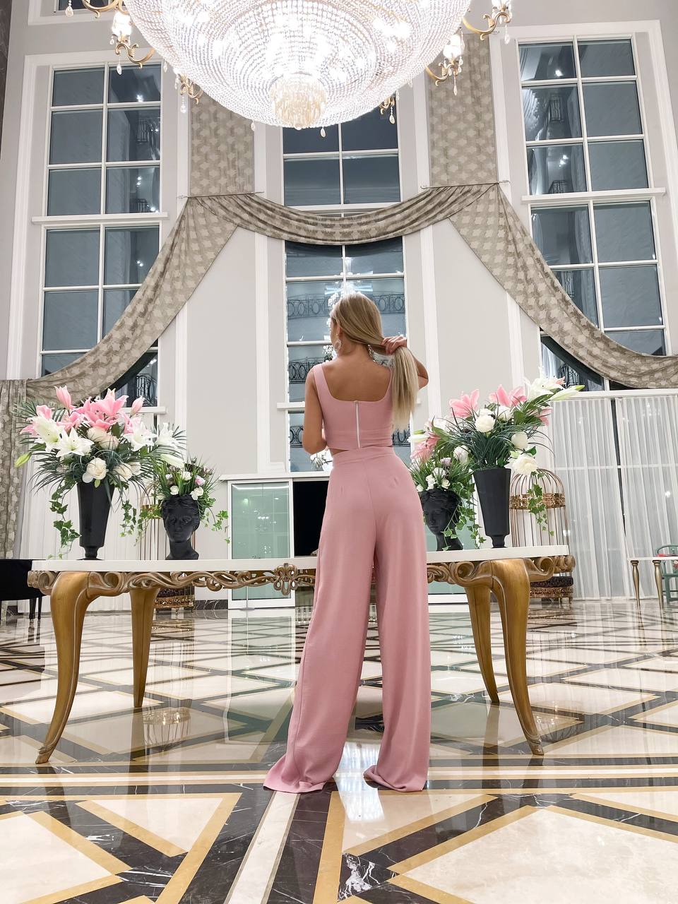 Woman in a pink jumpsuit standing in a elegant room with chandelier and floral arrangements.