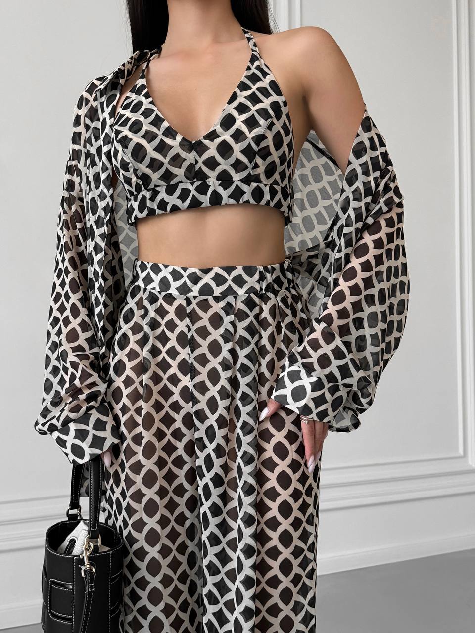 Woman wearing a patterned outfit with a black handbag against a white wall.