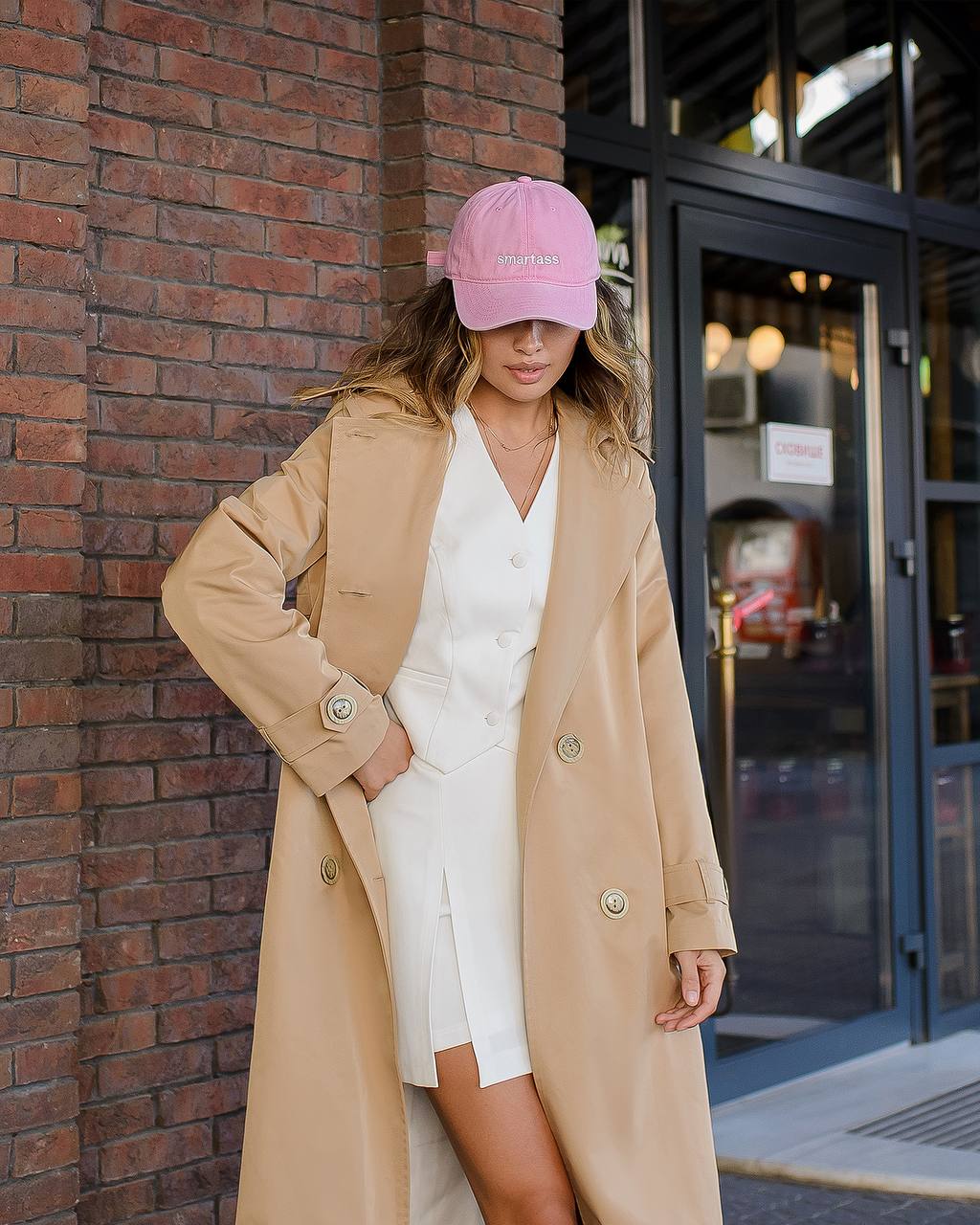Woman wearing a beige coat and pink cap leaning against a brick wall.