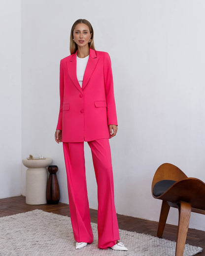 Woman wearing a bright pink suit standing in a minimalistic room with a wooden chair and white wall.