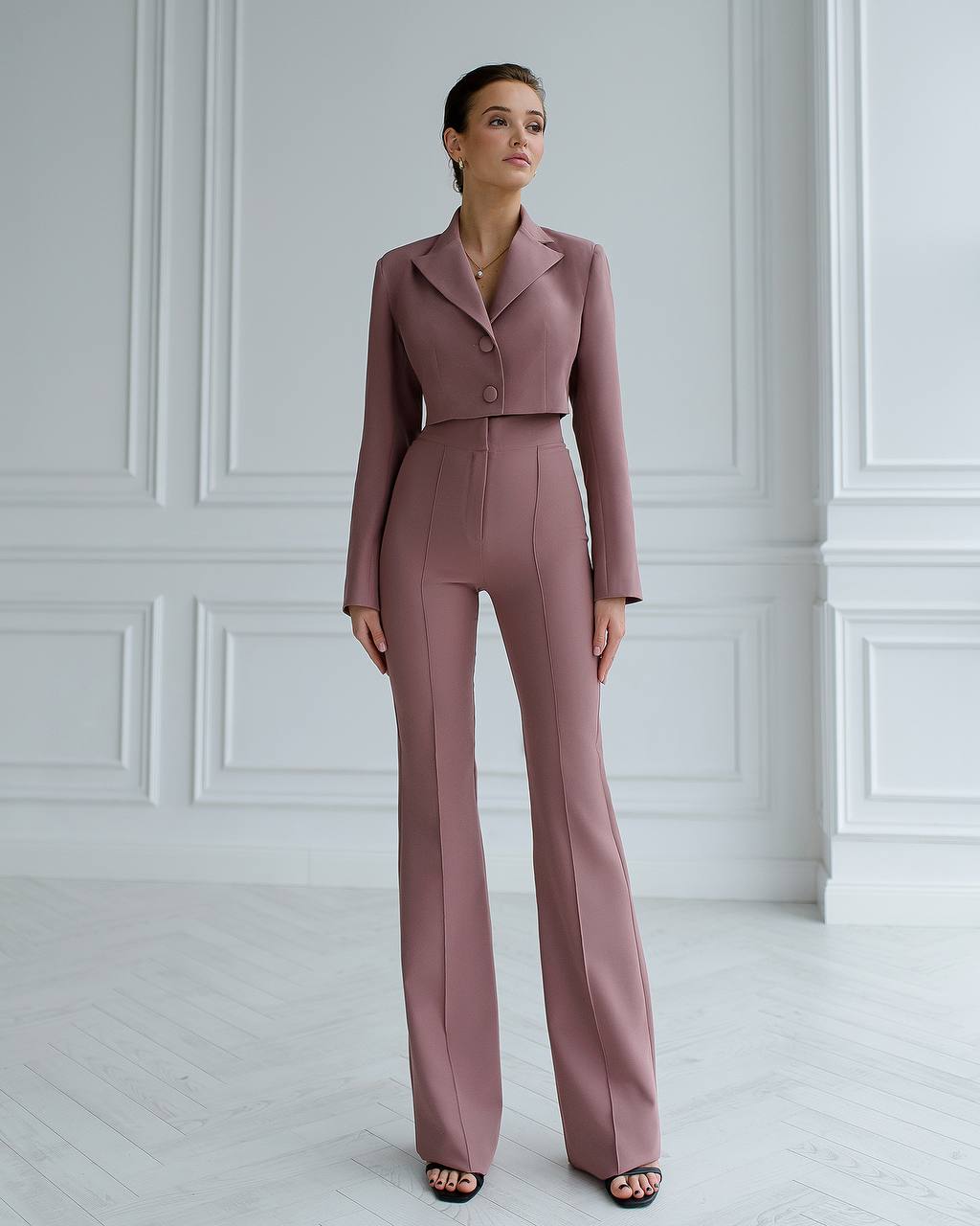 Woman wearing a mauve suit standing against a white paneled wall.